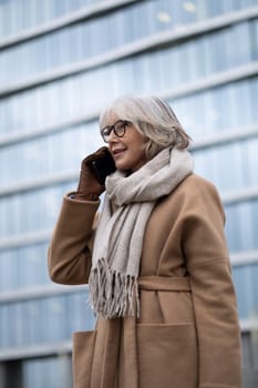 An older woman with gray hair happily chats on her phone, dressed warmly in a beige coat and scarf, in front of a sleek glass building reflecting sunlight.