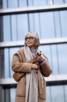 A senior woman dressed in a fashionable coat and scarf stands confidently outside a contemporary glass building. She holds a notebook while looking thoughtfully ahead.
