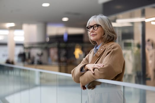 A woman with silver hair and glasses leans against a railing in a shopping mall, appearing contemplative as she looks around, surrounded by modern shops and people.