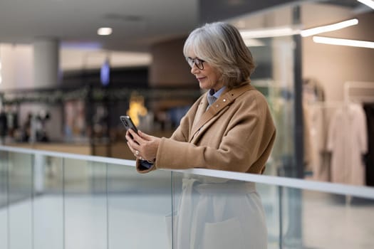 An older woman in a beige coat enjoys scrolling through her smartphone while leaning on a glass railing in a contemporary shopping mall. The atmosphere is bright and inviting.