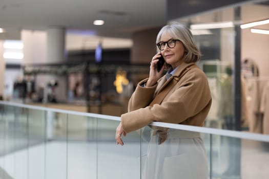 In a bustling shopping mall, a stylish mature woman with glasses engages in a phone conversation while casually leaning on a railing, showcasing a contemporary urban atmosphere.
