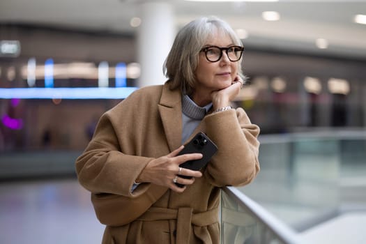 A woman with stylish glasses leans against a railing, thoughtfully holding her smartphone. She appears lost in contemplation in a contemporary setting filled with soft lighting.