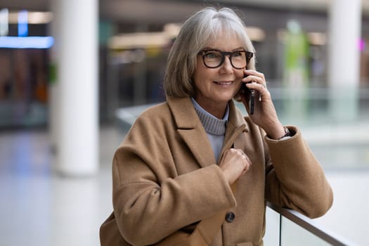 A cheerful older woman in a stylish beige coat talks on her mobile phone while standing by a railing in a contemporary shopping center filled with natural light.