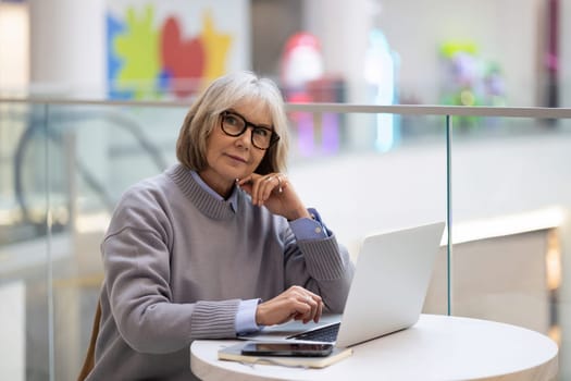 An older woman sits at a round table, thoughtfully working on her laptop in a bright and modern workspace. Creative decor surrounds her, adding to the vibrant atmosphere.
