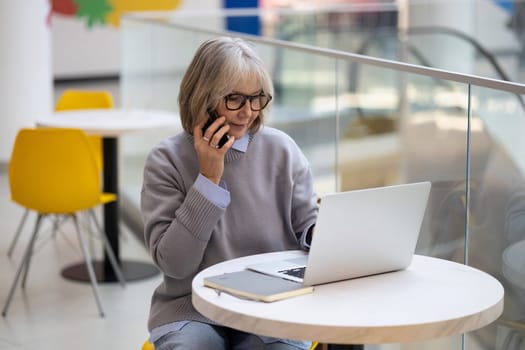 A senior woman engages in a phone conversation while focusing on her laptop in a bright, contemporary cafe. She appears concentrated and involved in her work.