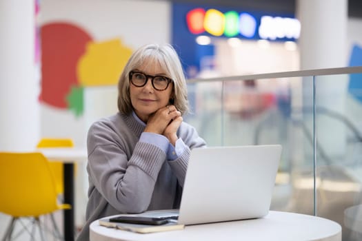 A senior woman with stylish glasses sits at a round table in a contemporary cafe. She works intently on her laptop, surrounded by colorful decor and bright lighting.