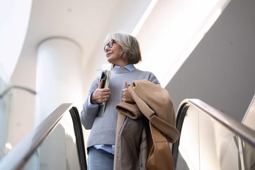 An older woman ascends an escalator in a contemporary building, holding a coat and a folder, deep in thought. Bright sunlight filters through large windows, creating a calm atmosphere.