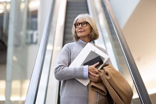A senior woman rides an escalator in a contemporary setting, carrying a tablet and brown coat. She appears thoughtful as she enjoys the ascent in a busy space.