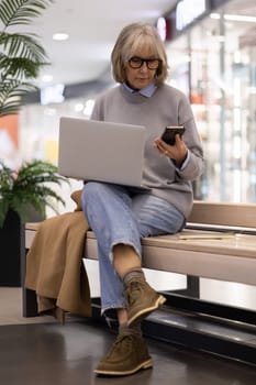 An older woman with glasses sits on a bench in a shopping mall, focused on her laptop and occasionally checking her smartphone, blending leisure with productivity.