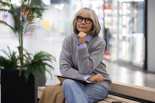 An older woman in a cozy gray sweater sits on a bench in a shopping mall, gazing thoughtfully while holding a notebook, surrounded by lush indoor plants, enjoying her day out.