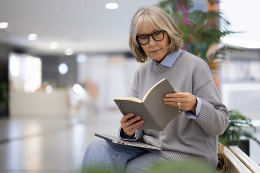 A woman with glasses sits comfortably on a bench, deeply engaged in reading a book. The surroundings showcase a contemporary indoor environment filled with plants and soft lighting.