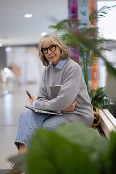 A woman with gray hair and glasses relaxes on a wooden bench, holding a notebook and pen while deep in thought, surrounded by indoor plants and a vibrant backdrop.
