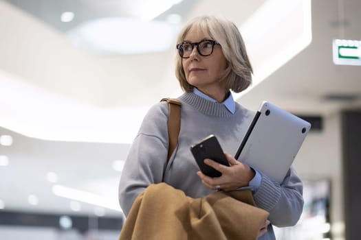 In a bustling shopping mall, a mature woman dressed in a sweater carries a laptop and smartphone, looking thoughtfully as she navigates the space during the afternoon.