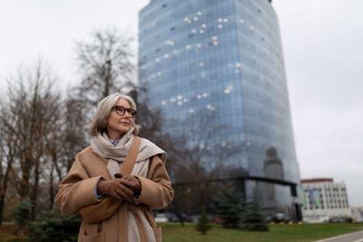 An elderly woman with stylish glasses smiles while standing outside in a brown coat and scarf. The backdrop features a contemporary glass building and bare trees under an overcast sky.