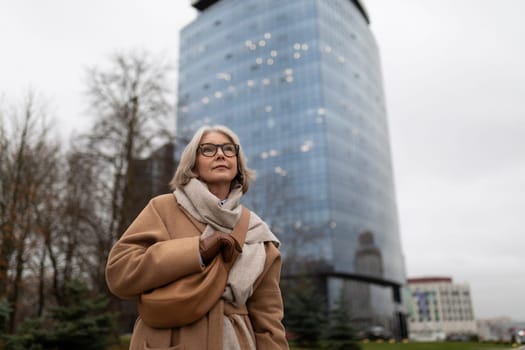 A senior woman with gray hair and glasses strolls through a city park, enjoying the fresh air. Behind her stands a contemporary glass skyscraper, reflecting the overcast sky.