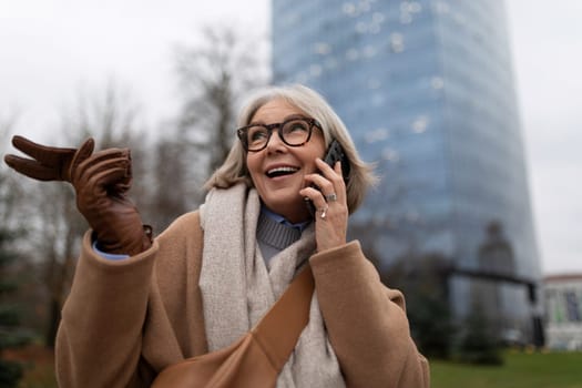 An older woman smiles brightly as she talks on her phone, gesturing joyfully with her hand while standing in a city park. The buildings loom in the background.