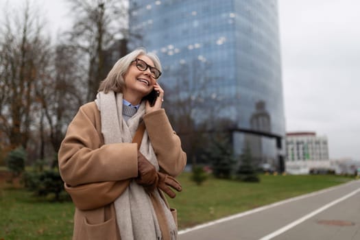 An elderly woman with glasses and gray hair smiles while talking on her phone. She wears a warm coat and gloves, surrounded by greenery and tall buildings.