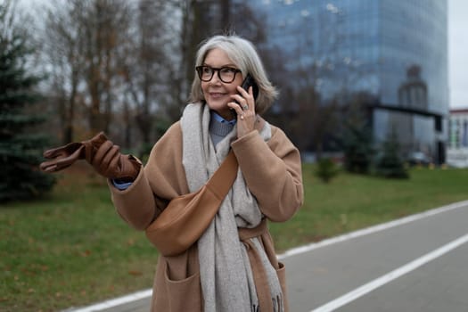 An older woman enjoys a stroll in the park, wrapped in a warm coat and scarf. She chats on her smartphone while holding a pair of gloves, surrounded by autumn scenery.