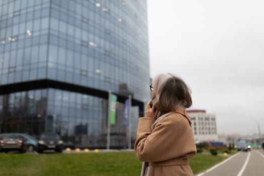 A woman with gray hair wearing a beige coat stands on a grassy area, engaged in a phone call next to a tall glass building on an overcast day in an urban setting.