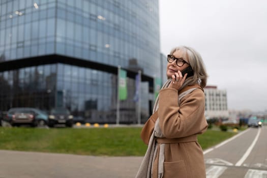 An older woman in a beige coat is engaged in a phone call, looking up thoughtfully while standing near a large glass office building. The weather is overcast and urban.