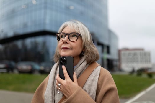 A mature woman dressed in a cozy coat and scarf stands outside, gazing upwards while holding her smartphone. The modern cityscape features glass buildings and overcast skies.