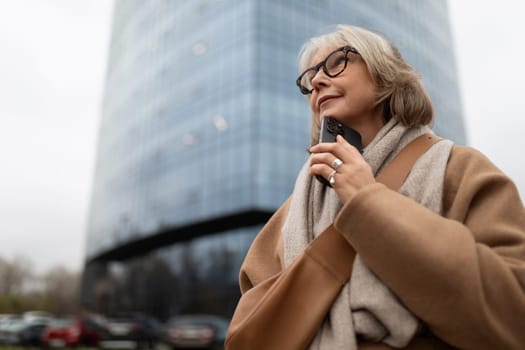 A thoughtful woman dressed warmly is holding a phone near her chin as she gazes upward, surrounded by a modern glass building under a cloudy sky.