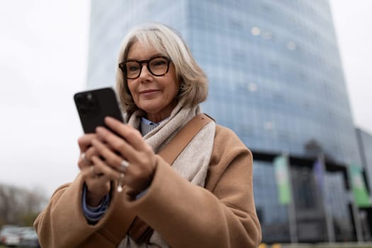 A woman with gray hair and glasses checks her smartphone while standing in front of a sleek, tall building. The weather is overcast, creating a calm atmosphere.