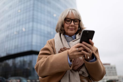 A woman dressed in a stylish coat and scarf is focused on her smartphone while standing in front of a contemporary glass building. The weather appears cool and overcast.