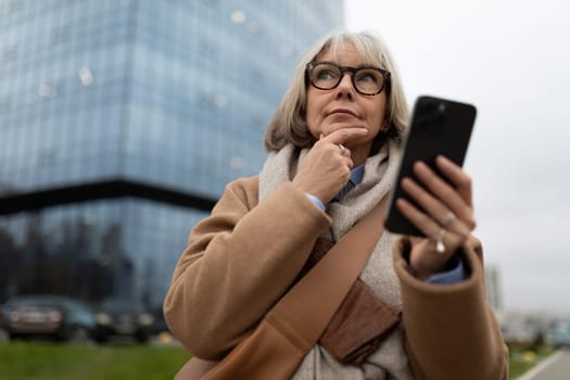 A senior woman stands outdoors, thoughtfully holding her smartphone. She appears to be considering something important, surrounded by a modern urban setting.