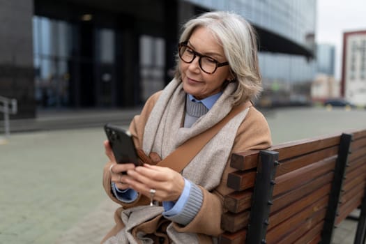 A woman with gray hair and glasses sits comfortably on a bench, attentively using her smartphone while enjoying the pleasant weather in the urban setting.