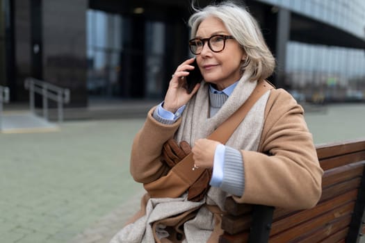 A senior woman dressed in a warm coat and scarf sits comfortably on a bench outside a modern office building. She is engaged in a conversation on her mobile phone, enjoying the sunny weather.