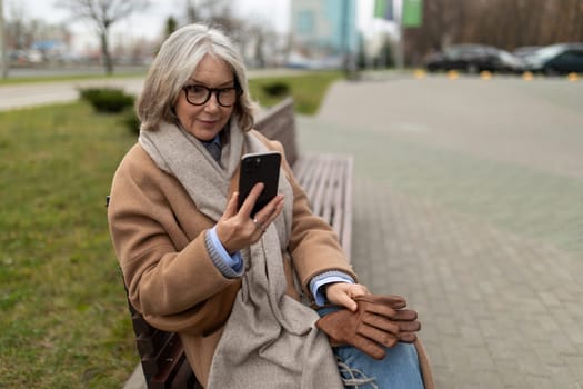 A middle-aged woman with gray hair and glasses dressed in a warm coat and scarf sits comfortably on a park bench. She is focused on her phone, enjoying a chilly afternoon.