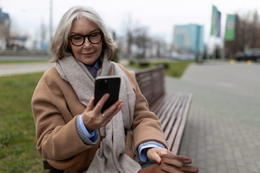 A woman with gray hair and glasses sits comfortably on a park bench, smiling as she interacts with her smartphone in a city environment during springtime.