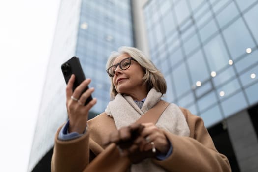 A mature woman stands outside a contemporary building, scrolling through her smartphone. She wears a warm coat and scarf, displaying a relaxed and engaged demeanor in an urban setting.