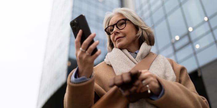 A mature woman wearing glasses and a warm coat is focused on her smartphone, standing on the sidewalk near modern buildings in an urban setting during overcast weather.