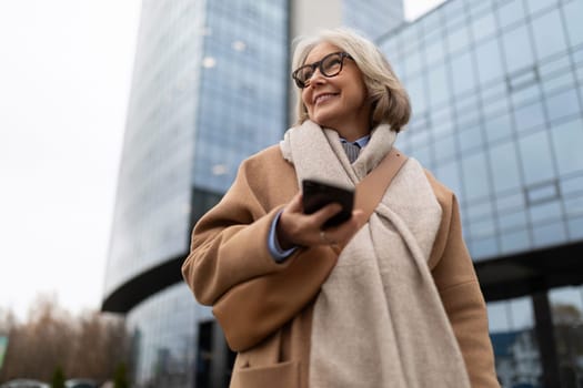 The woman, dressed in a warm coat and scarf, enjoys her time outdoors as she looks at her smartphone, surrounded by sleek, glass buildings during a cloudy afternoon.