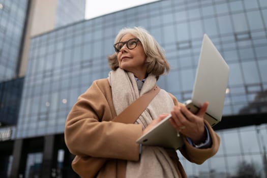 A mature woman stands outside a contemporary building, holding her laptop and looking thoughtfully into the distance. She wears a stylish coat and a cozy scarf, enjoying the moment.