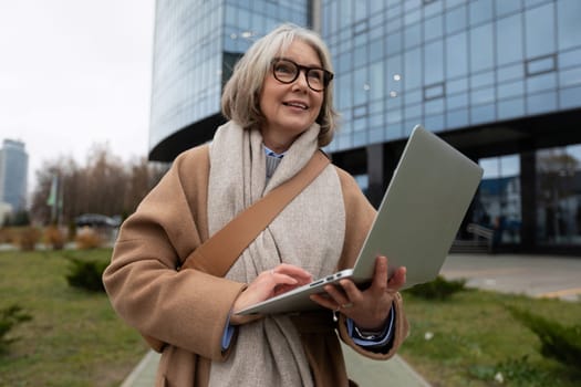A senior woman stands outside a contemporary office building, smiling as she types on her laptop. The setting reflects a blend of nature and urban life on a cool day.