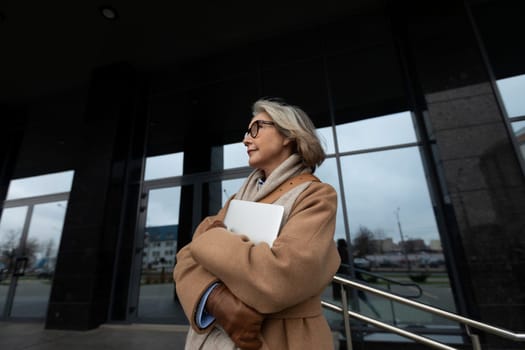 A woman with gray hair stands confidently outside a sleek, contemporary building. She is wearing a beige coat, holding a laptop, and looking pensively at the overcast sky.