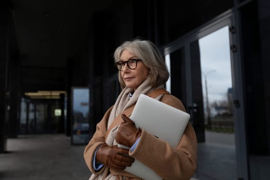 A stylish woman with gray hair stands outside a contemporary building. She holds a laptop while dressed warmly in a coat and scarf, reflecting a thoughtful expression.