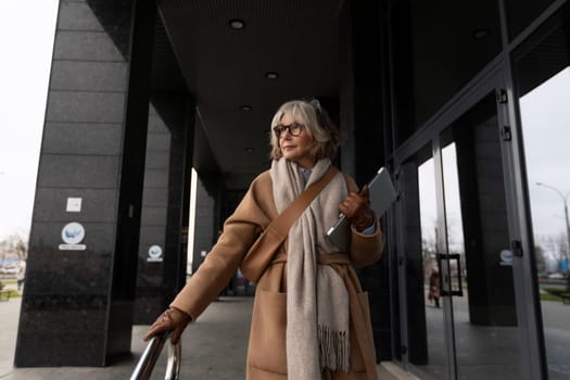 An older woman dressed in a fashionable coat and scarf steps out of a contemporary building, holding a tablet and enjoying the cool weather.