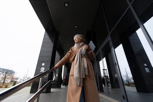 A woman wearing a fashionable coat and scarf descends a staircase outside a contemporary building. She holds a tablet in her hand, observing her surroundings on a cloudy day.