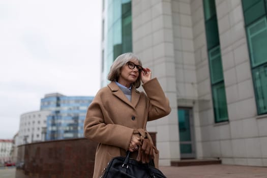 An older woman dressed in a stylish brown coat and gloves walks along a city street. She pauses to adjust her glasses while surrounded by contemporary architecture.