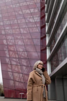 The woman walks confidently while speaking on her phone, dressed in a stylish beige coat and scarf. She stands beside a contemporary building with a geometric, purple exterior.