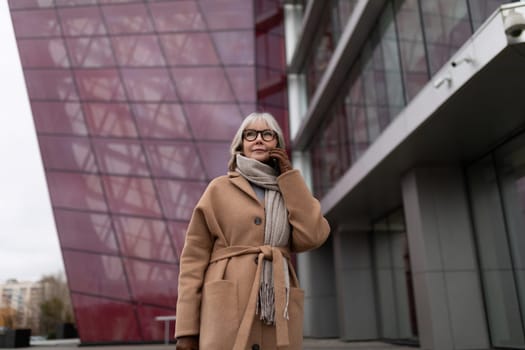 A woman with gray hair wearing glasses stands outdoors, looking thoughtfully while dressed in a cozy coat and scarf. The backdrop features modern architectural elements.