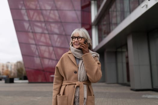 An elderly woman dressed in a warm coat and scarf smiles as she walks by a contemporary architectural structure on a cool, cloudy day in the city.