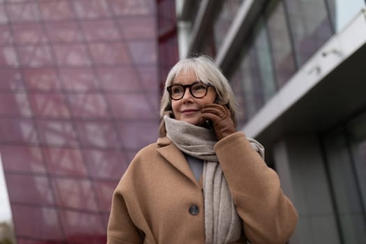A woman with gray hair, dressed in a warm brown coat and scarf, smiles while talking on her phone outside a contemporary building. It is daytime, and she appears content.