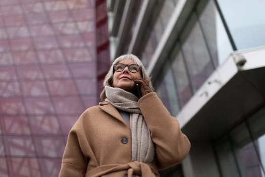 An elderly woman with glasses and a warm coat stands outside a contemporary urban building, engaged in a phone conversation. She appears confident and fashionable.