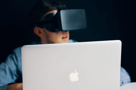 A young boy is seated in front of an Apple laptop, fully immersed in a virtual reality experience using a headset. Bishkek, Kyrgyzstan - March 14, 2020