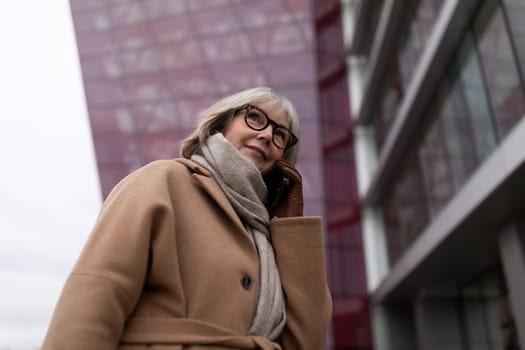 An older woman, wearing a tan coat and gray scarf, speaks on her smartphone while standing by a modern building with a colorful facade. The atmosphere is urban and overcast.